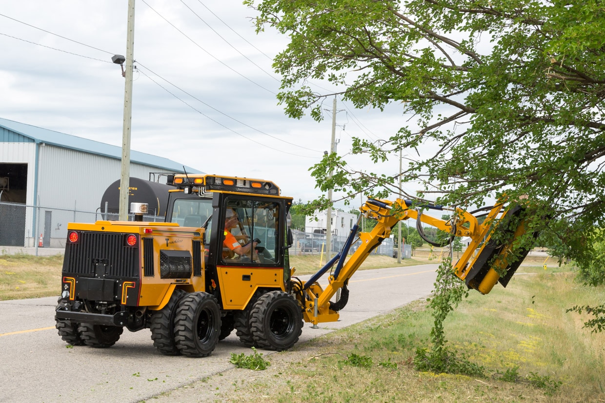 trackless vehicles boom flail mower attachment on sidewalk municipal tractor mowing grass in ditch rear side view