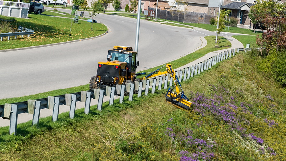 trackless vehicles boom flail mower attachment on sidewalk municipal tractor mowing grass in ditch rear view