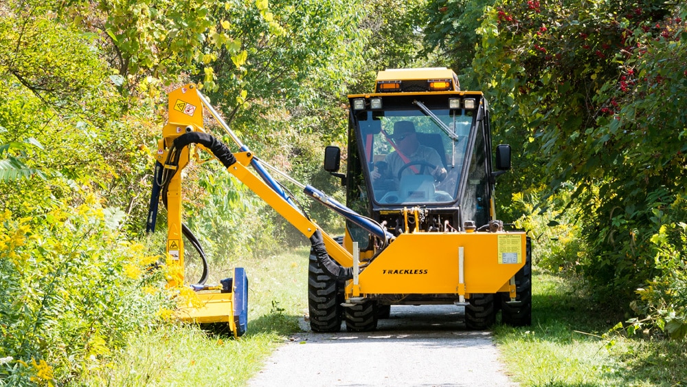 trackless vehicles boom flail mower attachment on sidewalk municipal tractor mowing grass front view